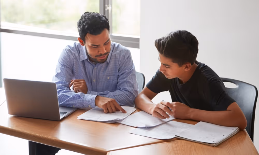 Tutor pointing at papers while helping a student sitting at a desk with a laptop and notebooks.