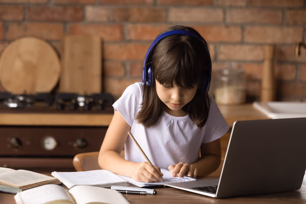 Young girl wearing headphones writing in notebook while using laptop at a table with open books.
