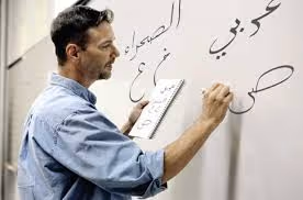 Man writing Arabic letters on a whiteboard while holding a notebook in his left hand.