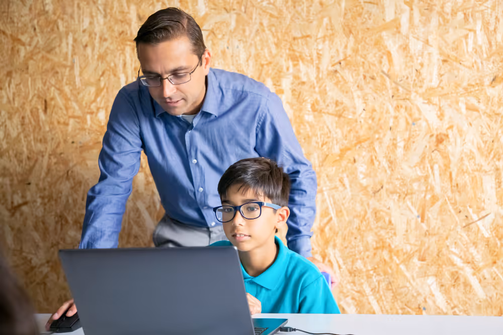Adult man in glasses helping a boy in glasses using a laptop at a table indoors.