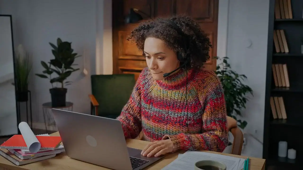 Person with curly hair and colorful sweater working on laptop wearing a headset in a cozy room.