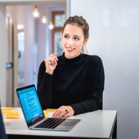 Eine lächelnde Frau, die mit einem Laptop und Unterlagen an einem Tisch sitzt.