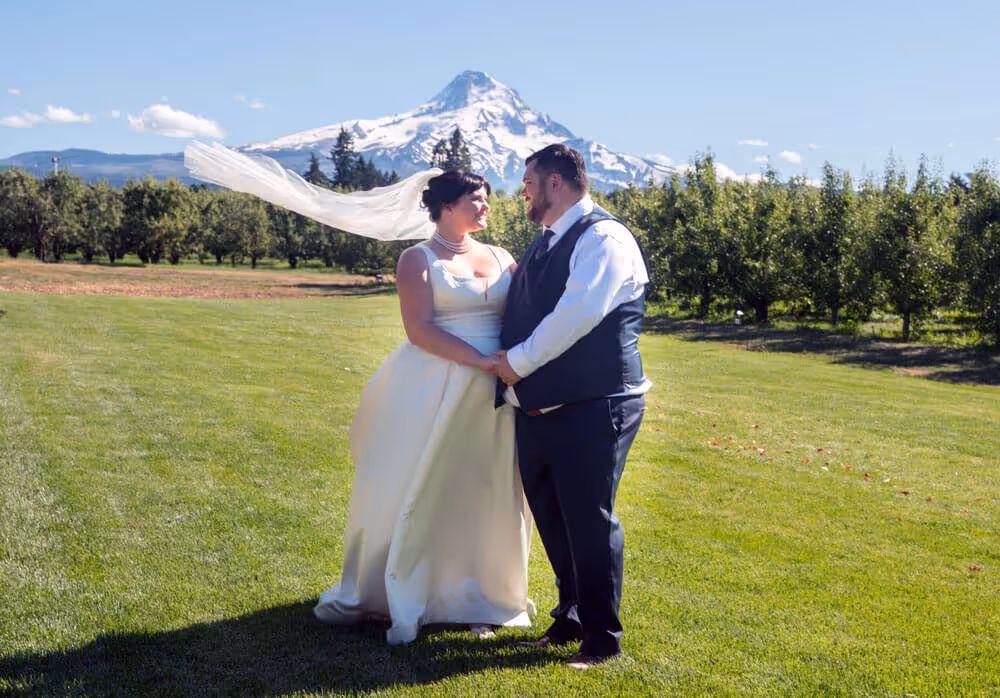 Bride in white dress and groom standing together on a grass lawn with Mt. Hood in the background