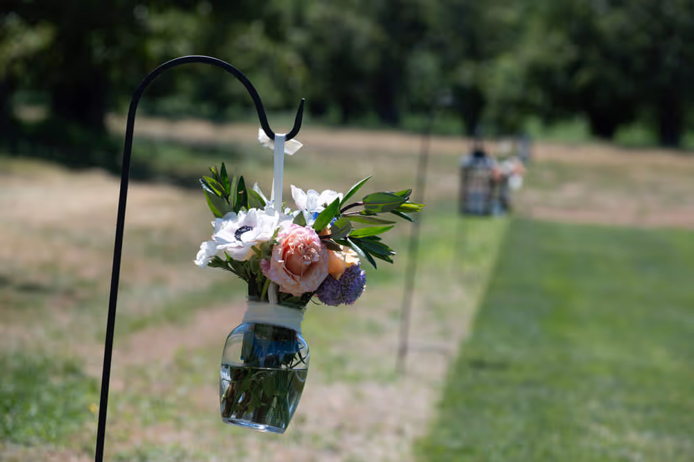 Small floral arrangement of roses and greenery in a glass jar hanging from a shepherd's hook ceremony decoration