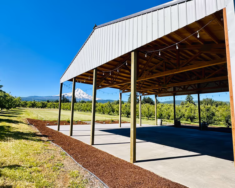 Open-air pole barn pavilion with concrete floor, surrounded by pear orchards and landscaped beds at The Perfect Pear.