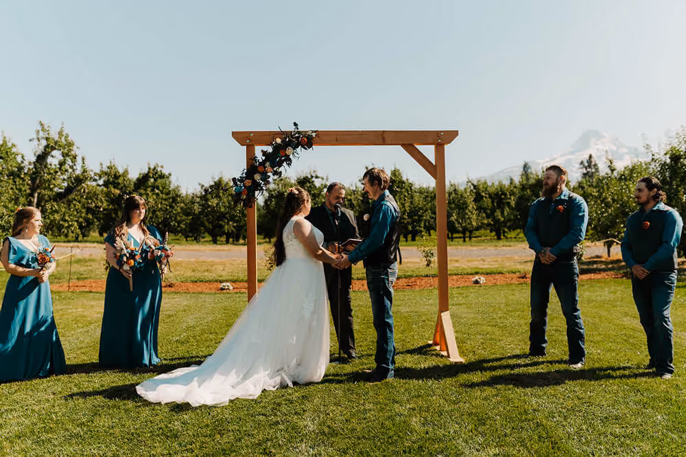 Couple holding hands under a wooden arch with bridesmaids and groomsmen on the orchard ceremony lawn, Mt. Hood behind them.