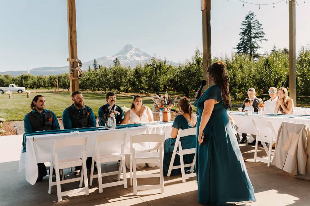 Bridesmaid giving a toast to the head table under the covered pavilion, with pear orchards and Mt. Hood in view.