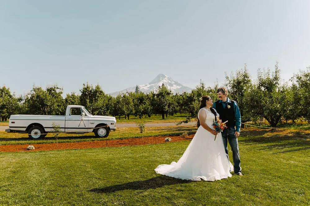 Bride and groom on the lawn beside a vintage white pickup truck, with pear trees and Mt. Hood in the background.
