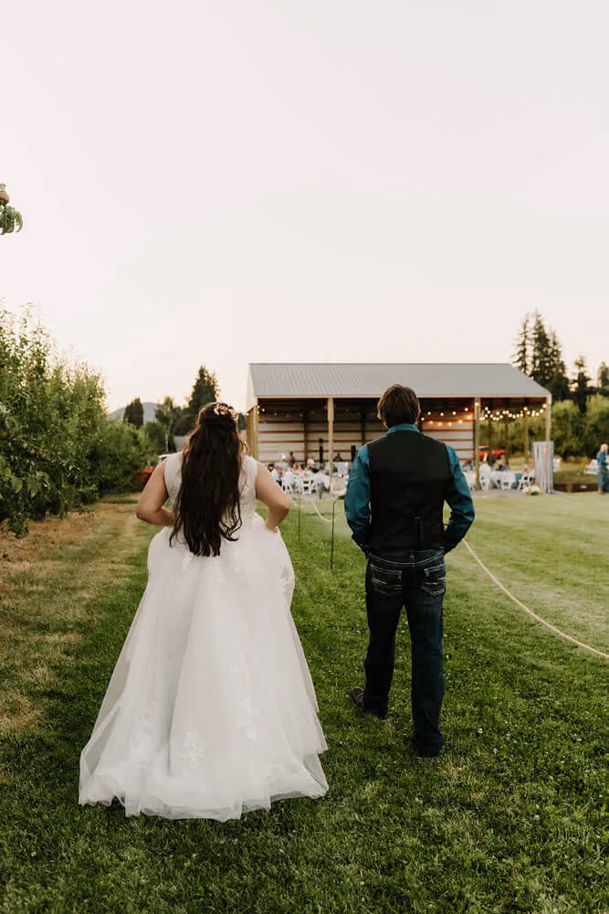 Bride and groom walking across the lawn at dusk toward the string-lit pavilion where their reception is underway.