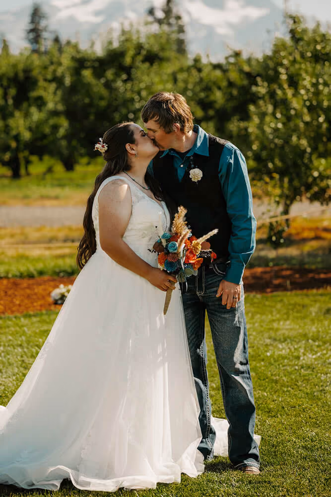 Groom kissing the bride on the lawn as she holds an orange and teal bouquet, with rows of pear trees behind them.