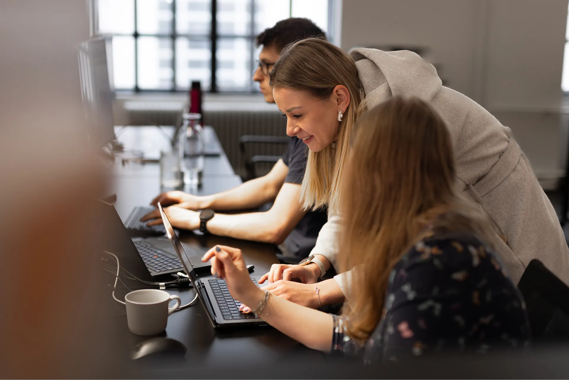 Zwei Frauen arbeiten gemeinsam an einem Laptop in einem hellen Büro, während ein Mann im Hintergrund am Computer arbeitet.