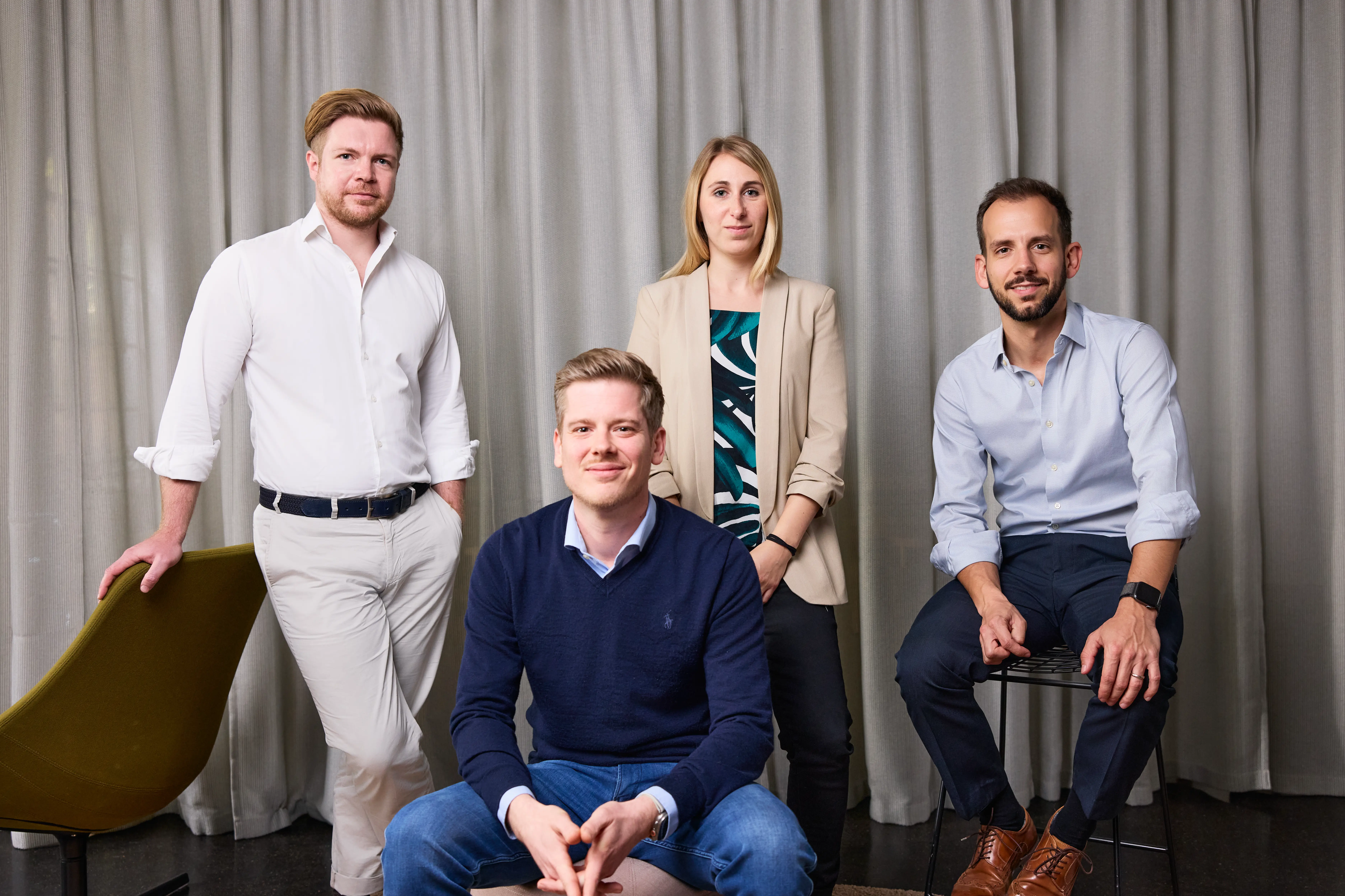 Vier professionelle Erwachsene, zwei Männer im Vordergrund sitzen, zwei Personen im Hintergrund stehen vor einem grauen Vorhang.