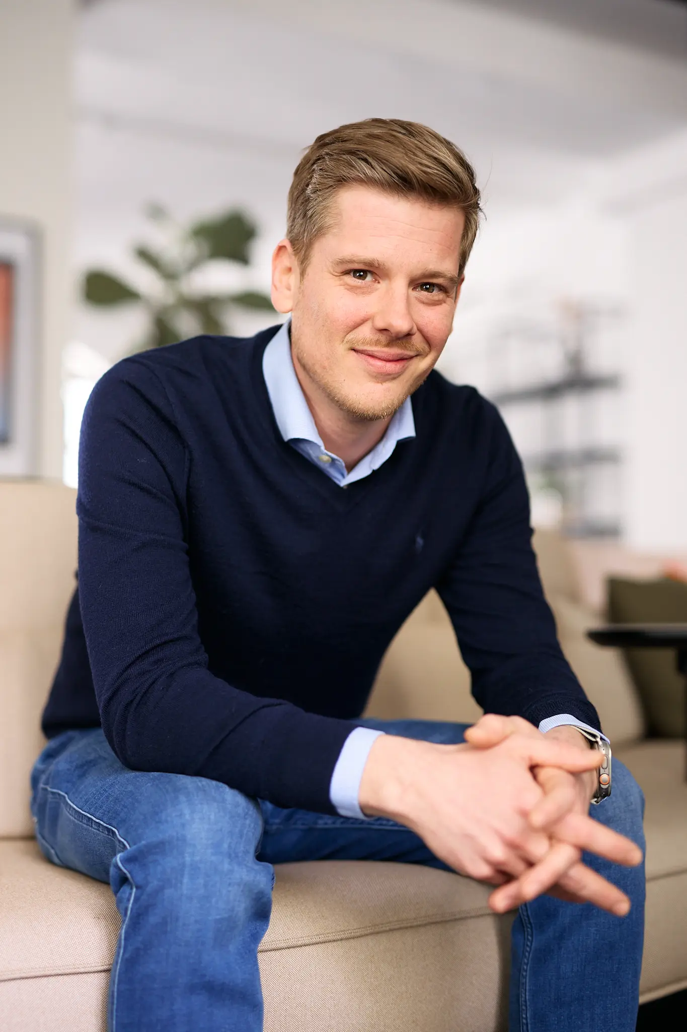 Junger Mann mit blauem Pullover und Hemd sitzt lächelnd auf einem Sofa in einem hellen Wohnzimmer.