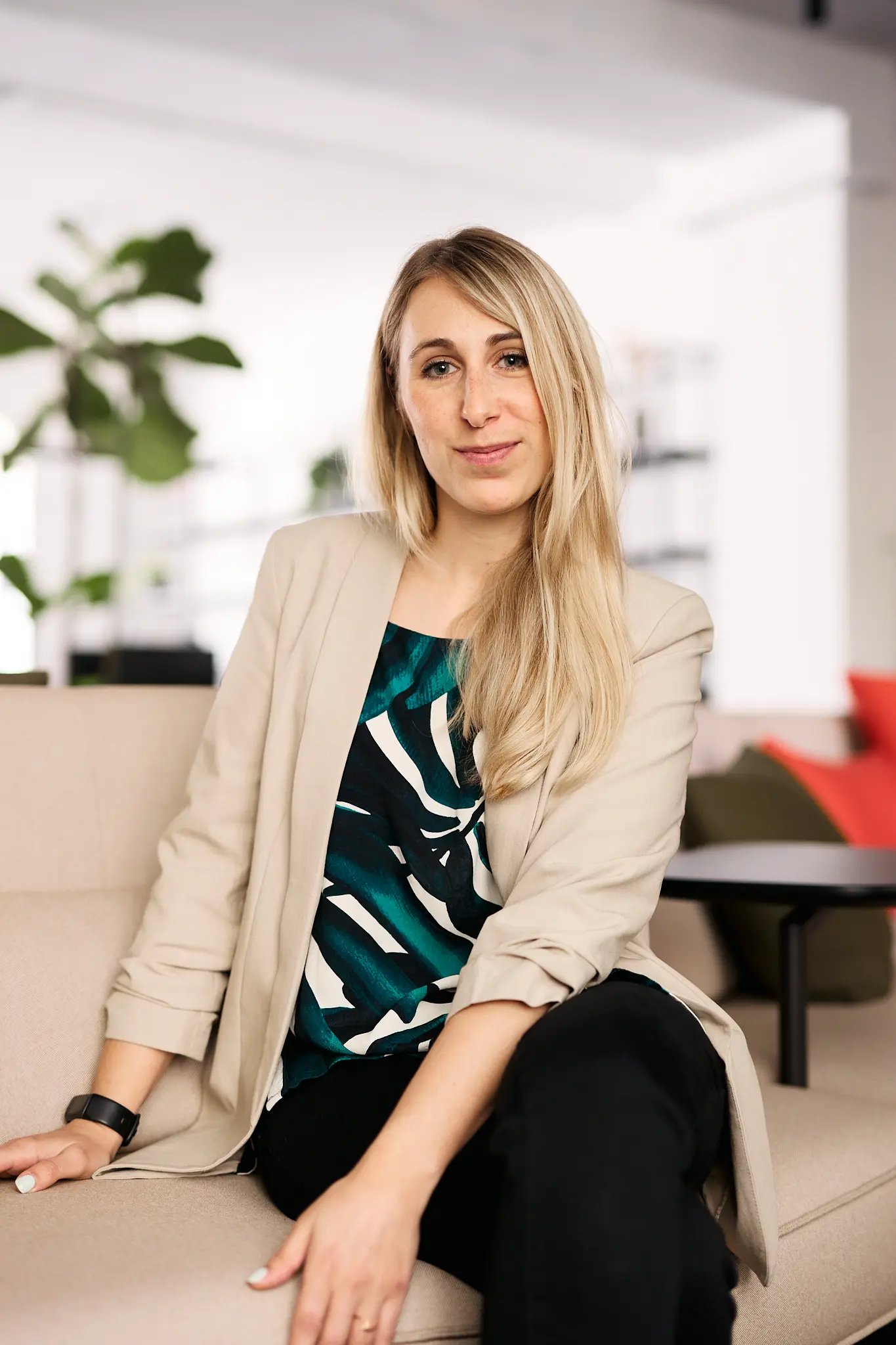 Frau mit langem blondem Haar, die in einem beige Blazer und gemustertem Oberteil auf einem Sofa sitzt.
