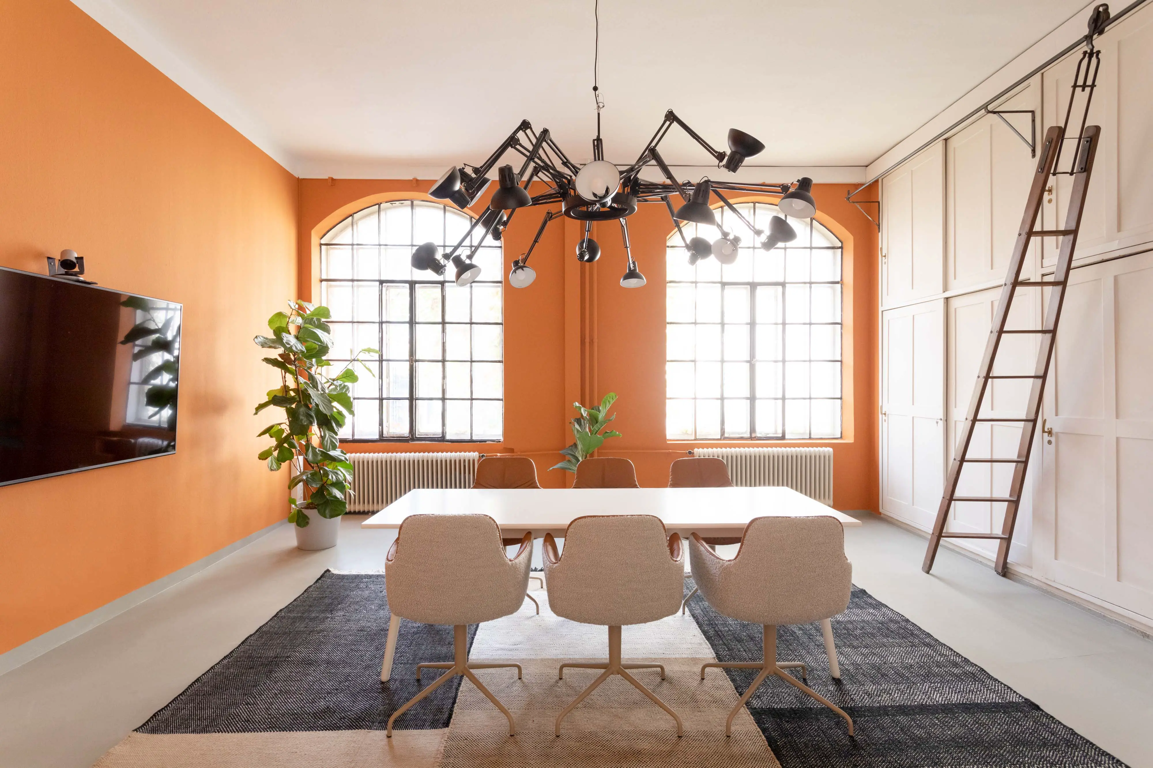 Moderner Besprechungsraum mit orangefarbenen Wänden, großem weißen Tisch, sechs gepolsterten Stühlen, großen Fenstern, hängender Mehrarm-Leuchte, einem großen Fernseher, Pflanzen und einer Leiter an weißen Schranktüren.