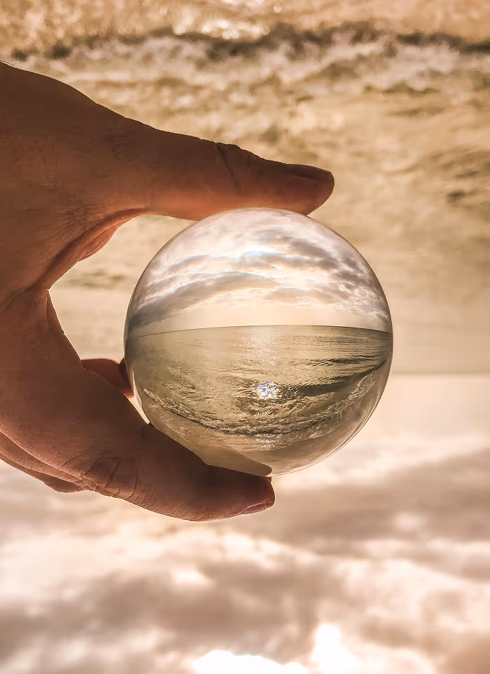 Eine Hand hält eine Glaskugel, in der sich ein spiegelverkehrtes Bild vom Meer und bewölktem Himmel zeigt.