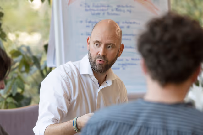 Stefan Puppe mit weißem Hemd führt eine Gesprächsrunde vor einem Flipchart mit Notizen, zwei Personen sind im Vordergrund unscharf zu sehen.