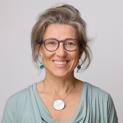 Anne Franko lächelt und trägt eine Brille, türkisfarbene Ohrringe und ein mintgrünes Oberteil.