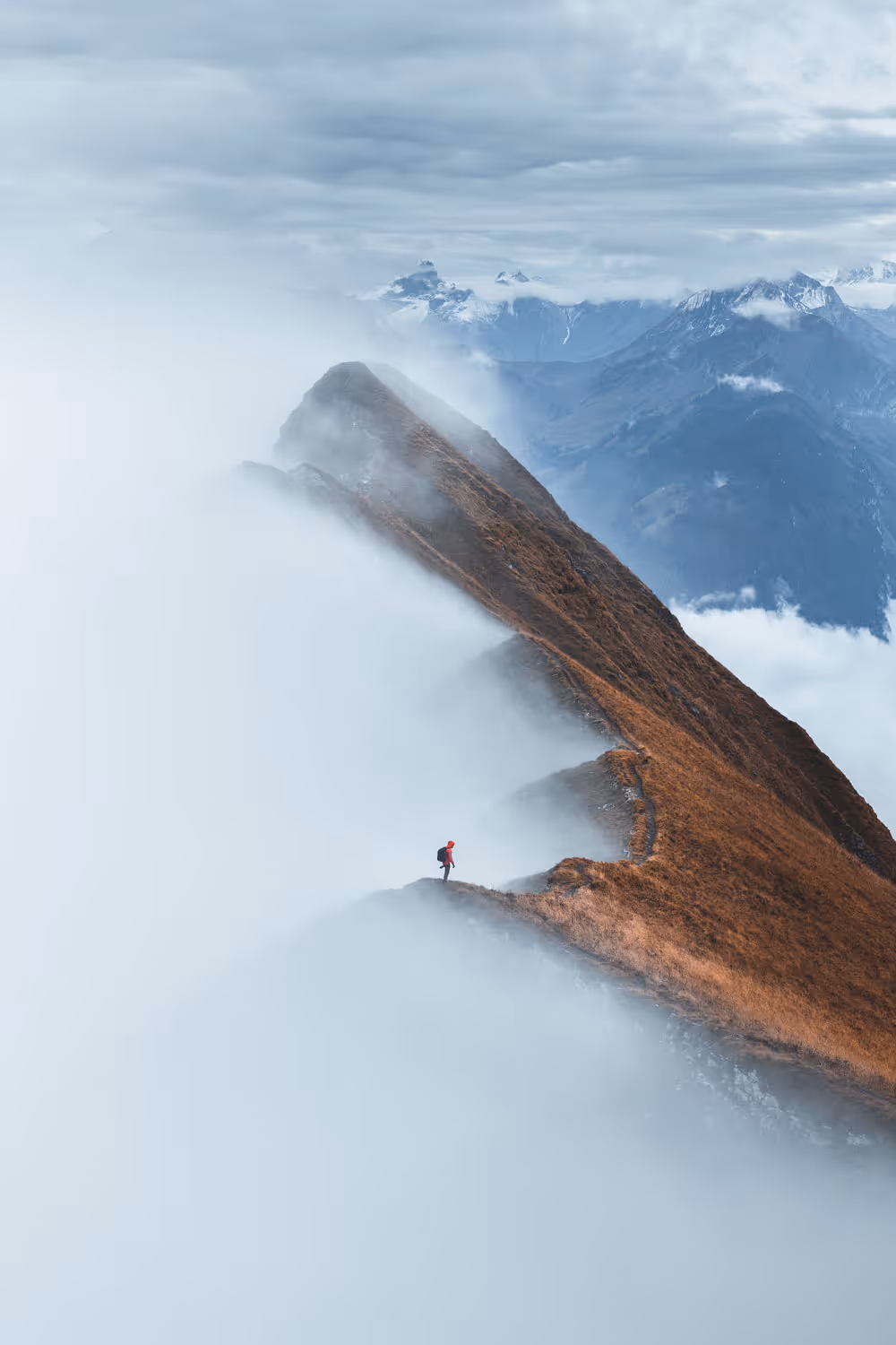 Wanderer in roter Jacke steht auf nebligem Berggrat mit Blick auf entfernte schneebedeckte Berge.