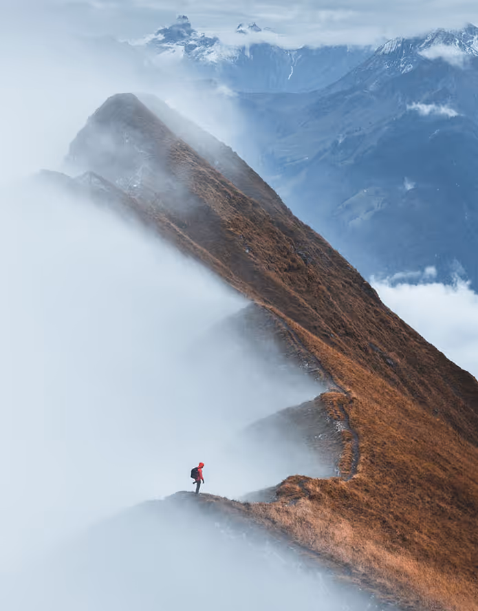Wanderer in roter Jacke auf einem Bergpfad umgeben von Nebel und Bergen im Hintergrund.