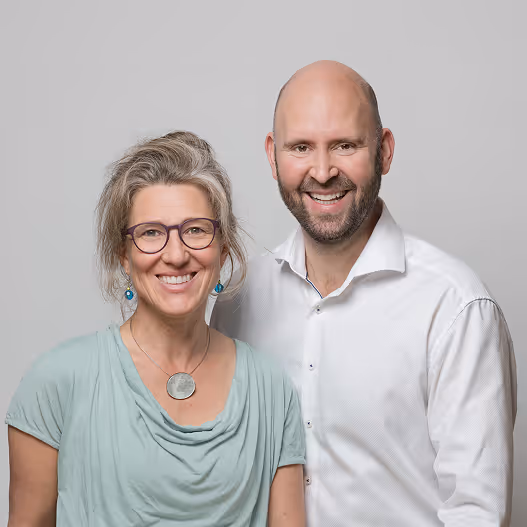 Zwei lächelnde Personen stehen vor grauem Hintergrund, Frau mit Brille, Mann mit Bart und weißem Hemd.