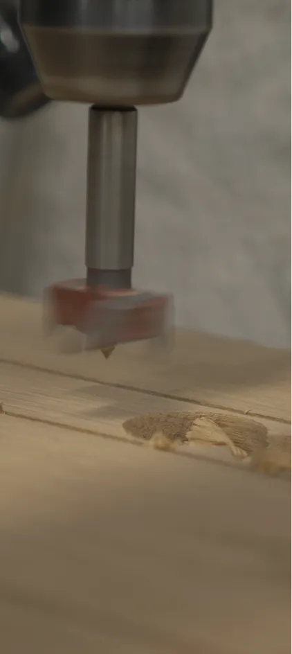 Close-up of a drill bit cutting a hole into a wooden board with wood shavings scattered.