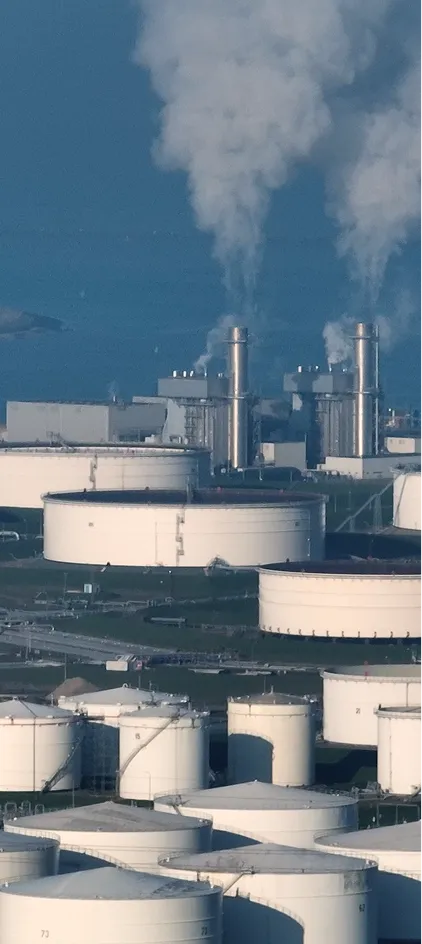 Industrial oil storage tanks with smokestacks emitting large plumes of smoke against a blue sky.