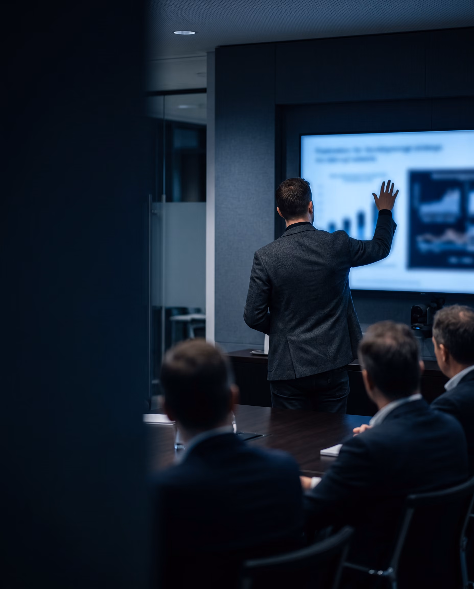 Man in a suit presenting data on a large screen to an audience of three men seated at a conference table in a modern office.