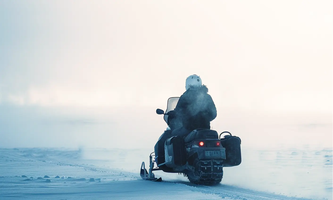 Person wearing a helmet riding a snowmobile on a snowy landscape with mist or fog in the background.