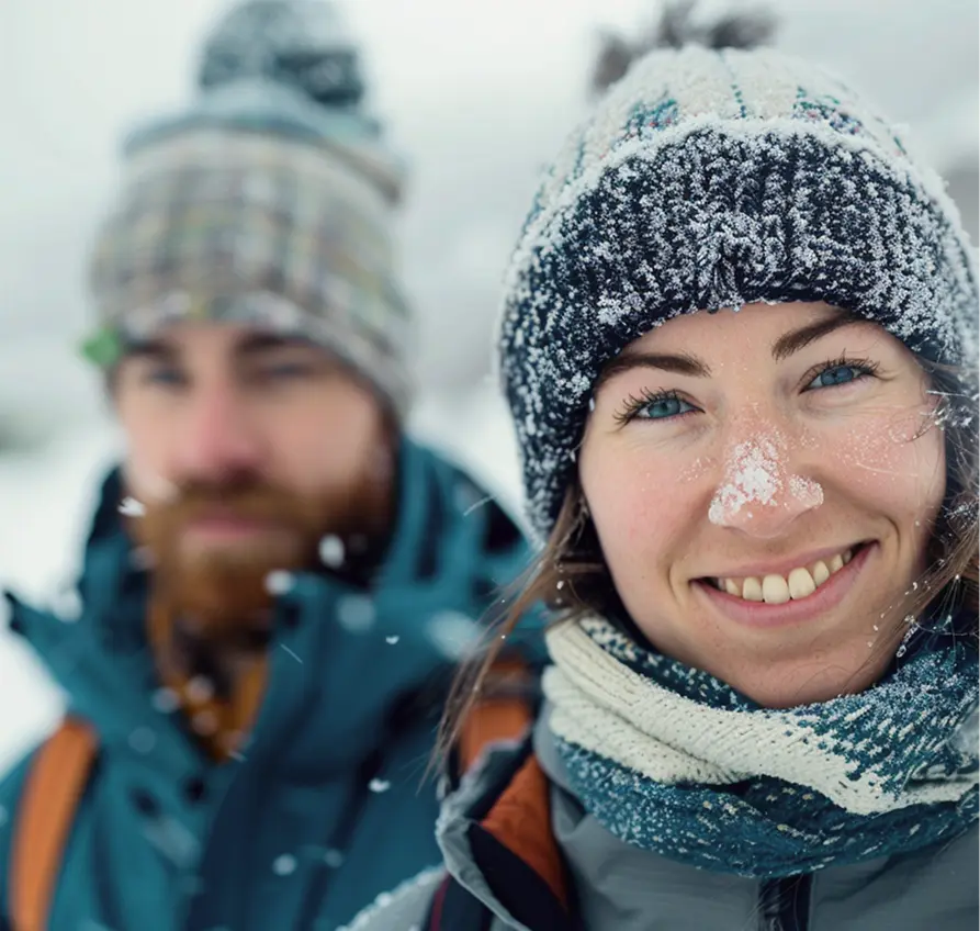 Smiling woman with snow on her nose wearing a winter hat and scarf, with a man in winter clothes blurred in the background.
