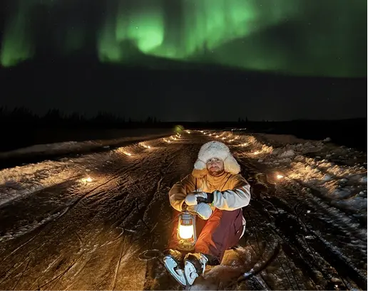 Person dressed in winter clothing sitting on a snowy path at night holding a lantern, with green northern lights glowing in the sky above.