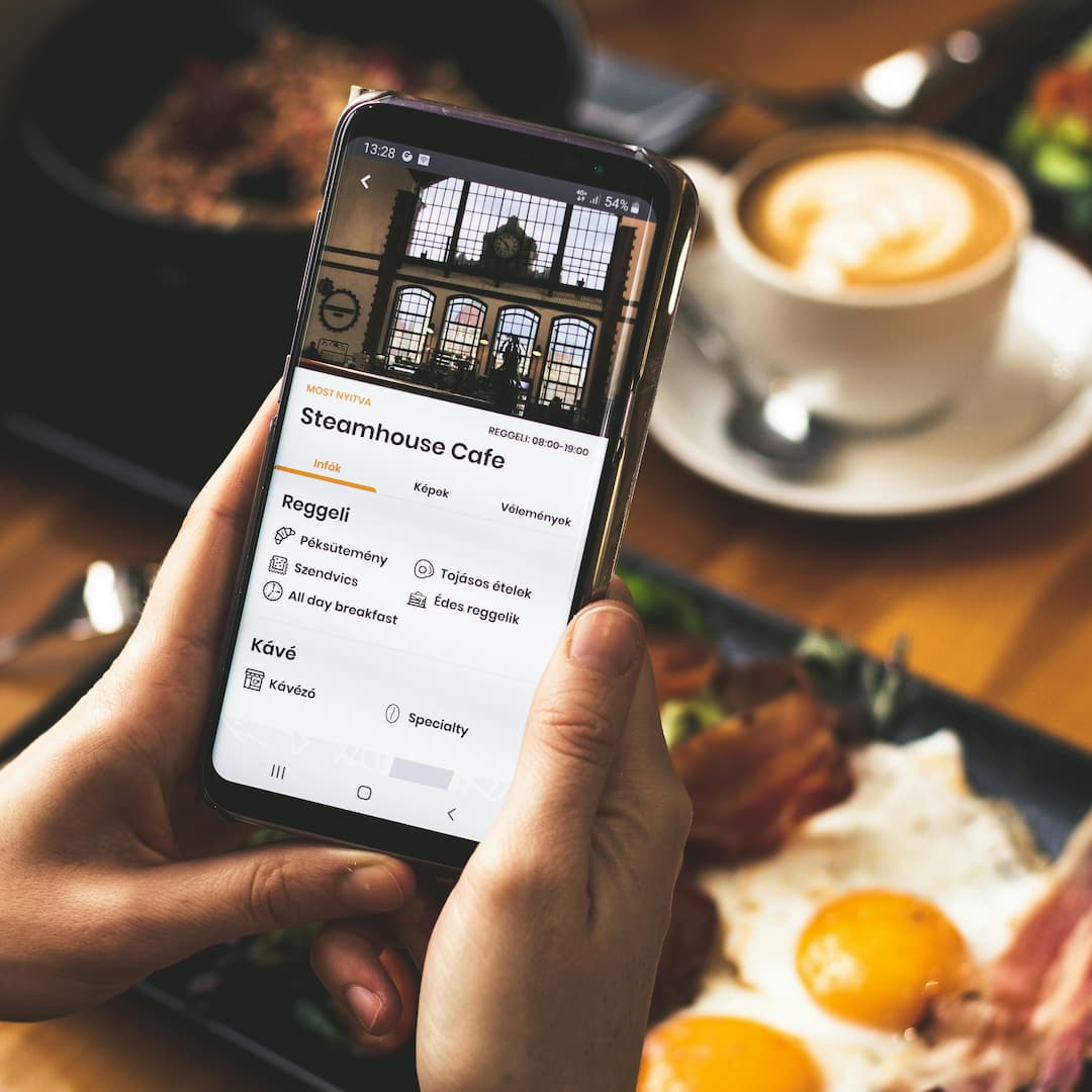 Une personne tenant un téléphone affichant la page du Steamhouse Cafe avec un menu de petit-déjeuner, à côté d'une assiette avec des œufs au plat et du bacon, et une tasse de café latte.