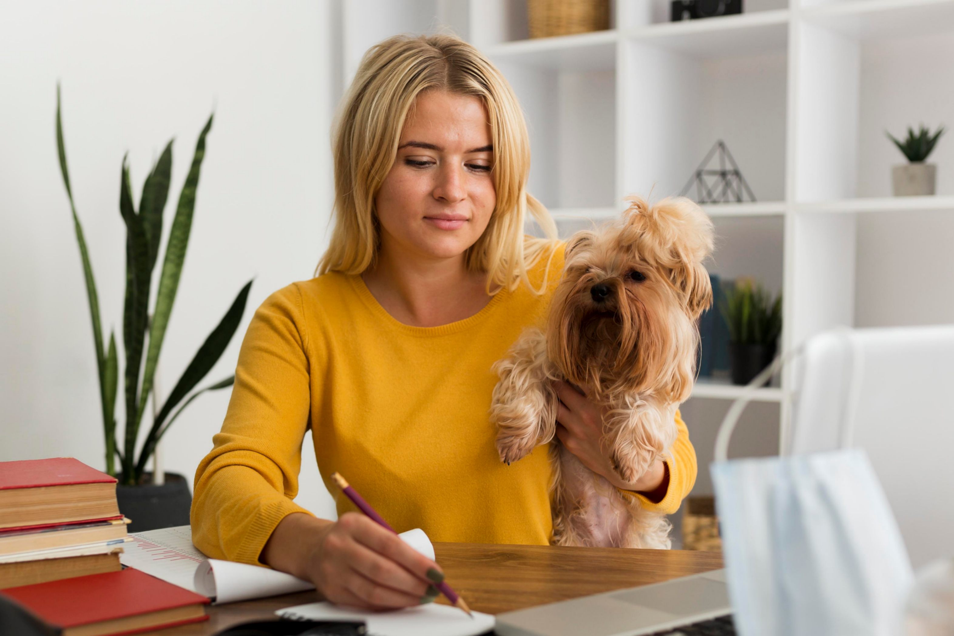 Une femme au pull jaune écrit dans son bloc note tenant un chien sous le bras