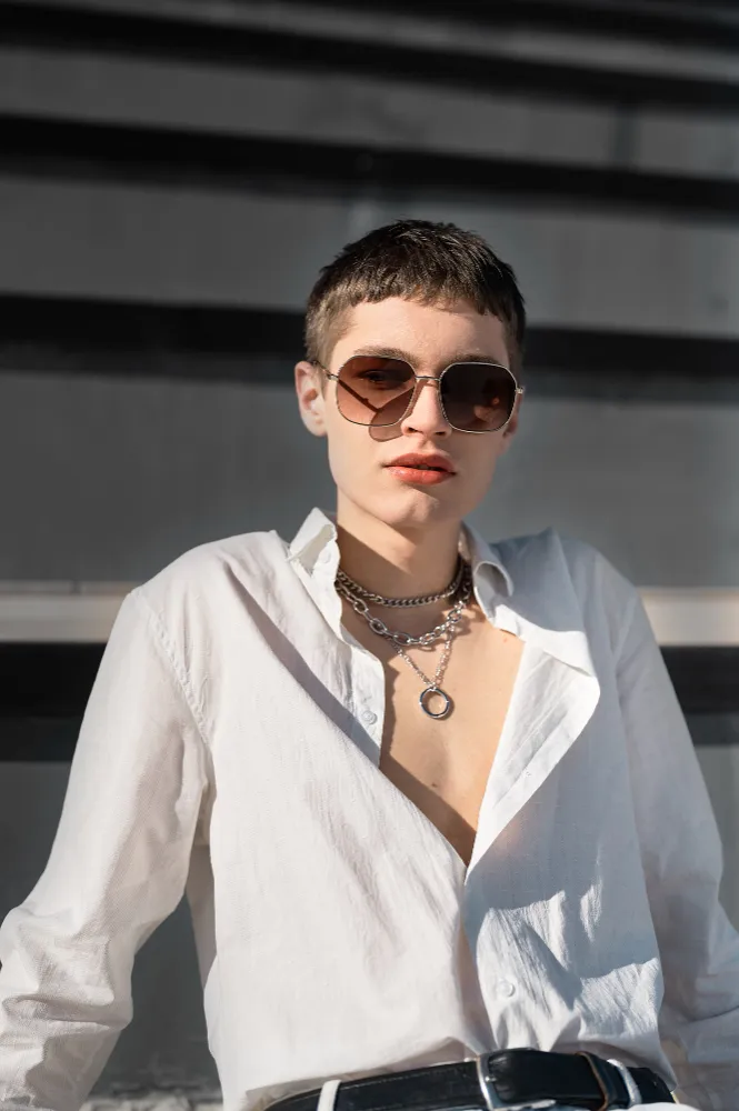 Person wearing brown sunglasses, layered silver chain necklaces, and white open shirt against dark background.