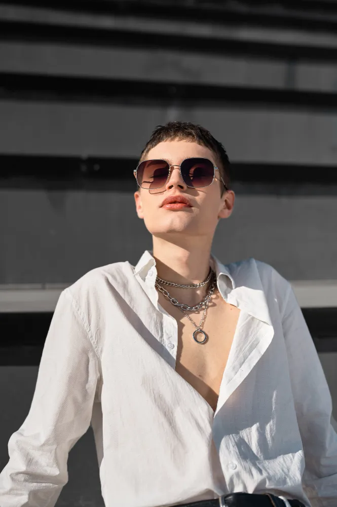 Person wearing sunglasses, white shirt with open collar, and layered silver chain necklaces against a dark background.