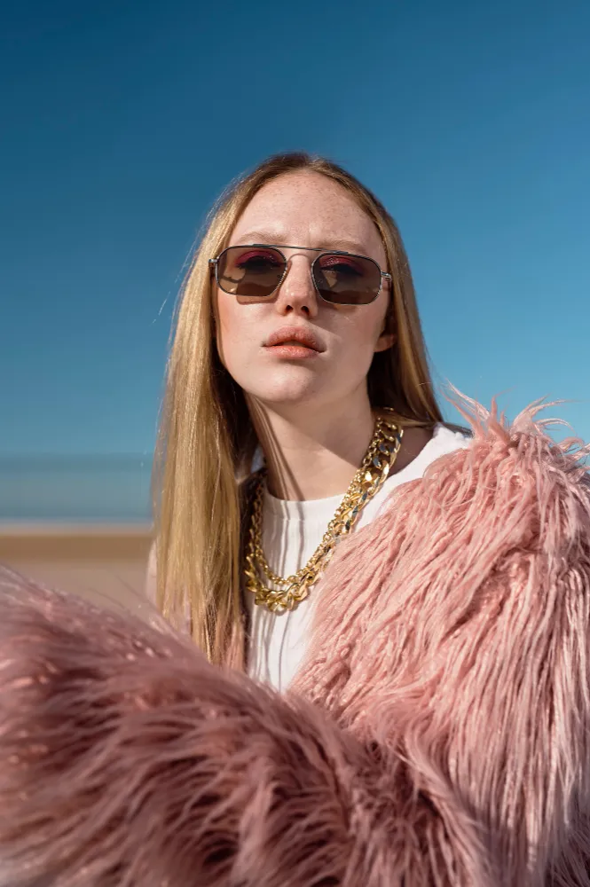 Young woman wearing sunglasses, gold chain, and pink furry jacket against clear blue sky.