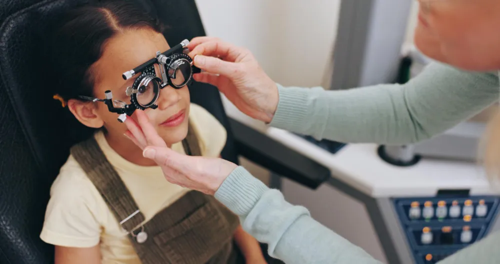 Child sitting in an eye exam chair while an optometrist adjusts trial frame glasses on their face.