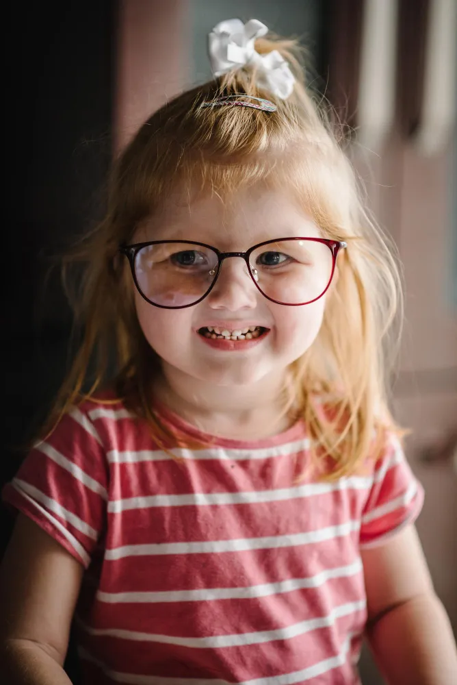 Smiling young girl with glasses, a white hair bow, and a red striped shirt.