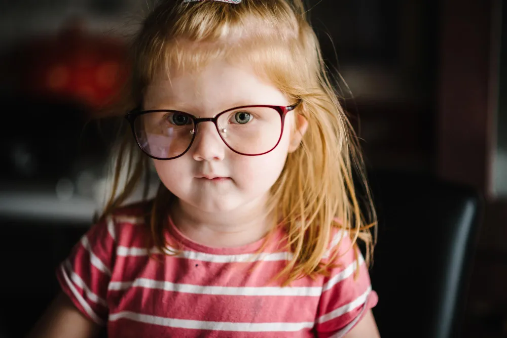 Young child with light brown hair wearing large glasses and a pink striped shirt looking at the camera.