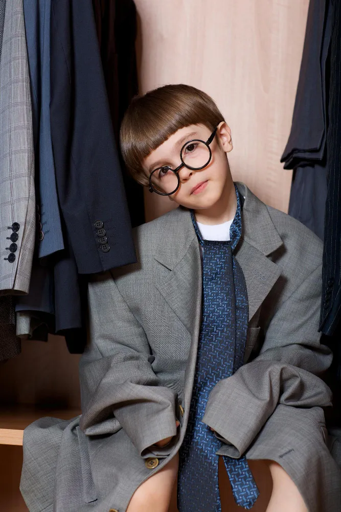 Young boy with round glasses wearing an oversized gray suit and blue tie sitting in a closet.