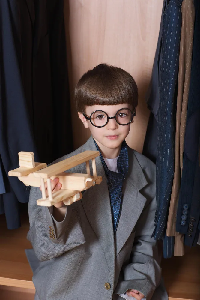 Child wearing oversized suit and round glasses holding a wooden toy airplane inside a closet.