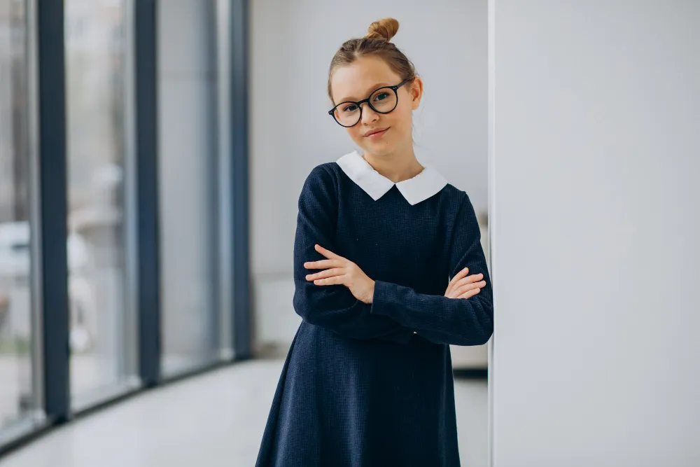 Young girl in glasses and dark dress with white collar standing with arms crossed by a wall.