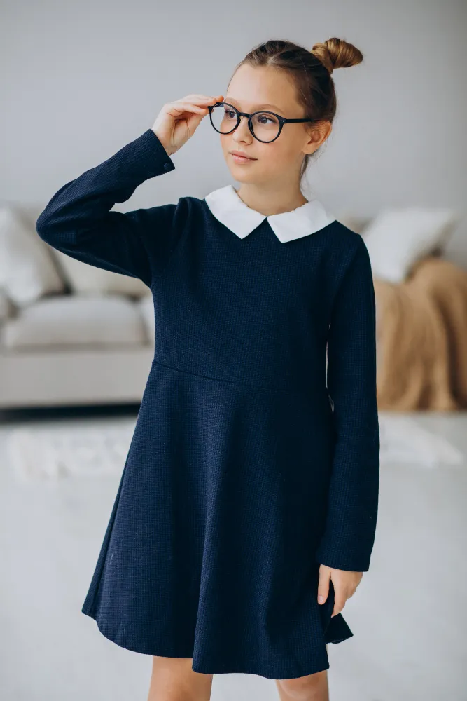 Young girl with glasses wearing a navy dress with white collar standing indoors.