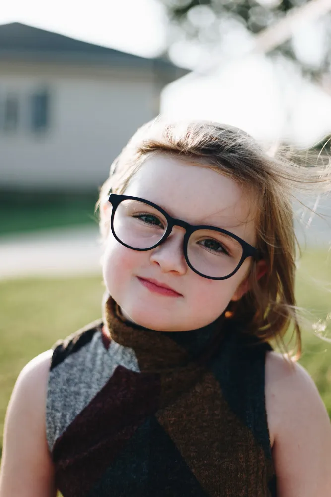 Young child with glasses, light brown hair, and a sleeveless patterned top outdoors on a sunny day.