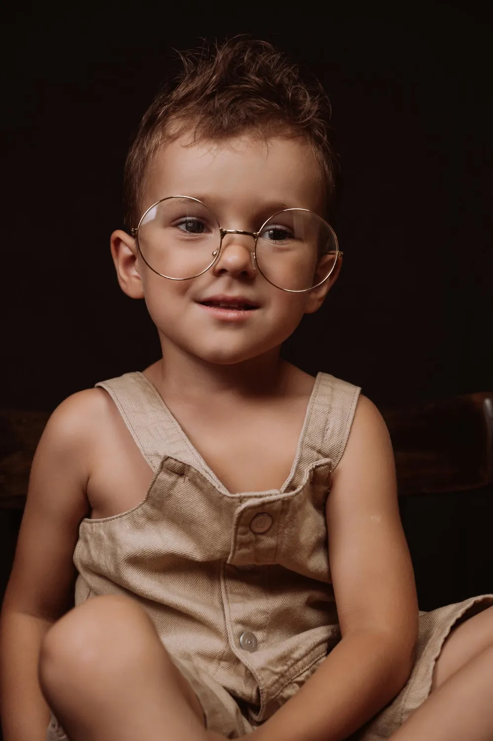 Young boy with short hair wearing large round glasses and a beige sleeveless outfit sitting against dark background.