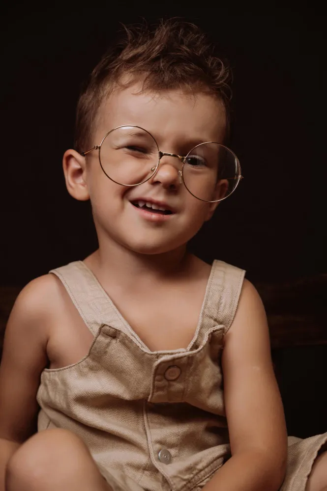 Young boy with short hair wearing large round glasses and beige overalls smiling with one eye closed.