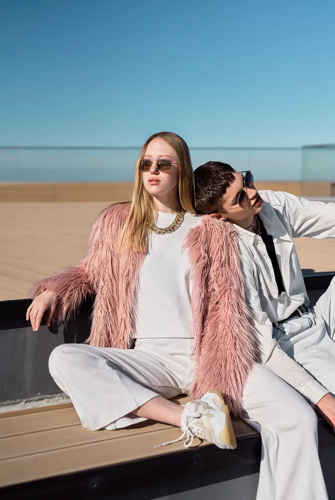 Two young people in sunglasses sitting outdoors on a wooden deck under clear blue sky.