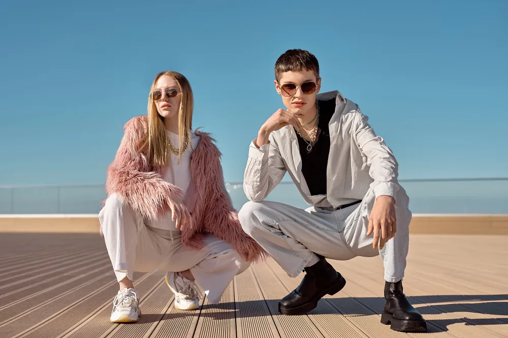 Two fashionably dressed people crouching on a wooden deck under clear blue sky wearing sunglasses.