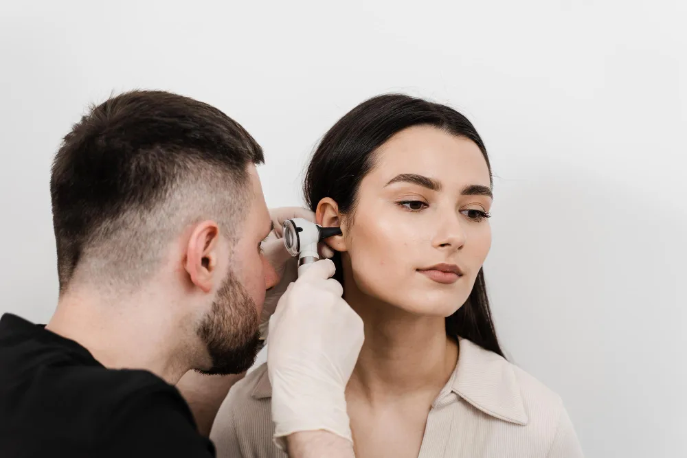 Man in gloves examining a woman’s ear with an otoscope against a plain white background.
