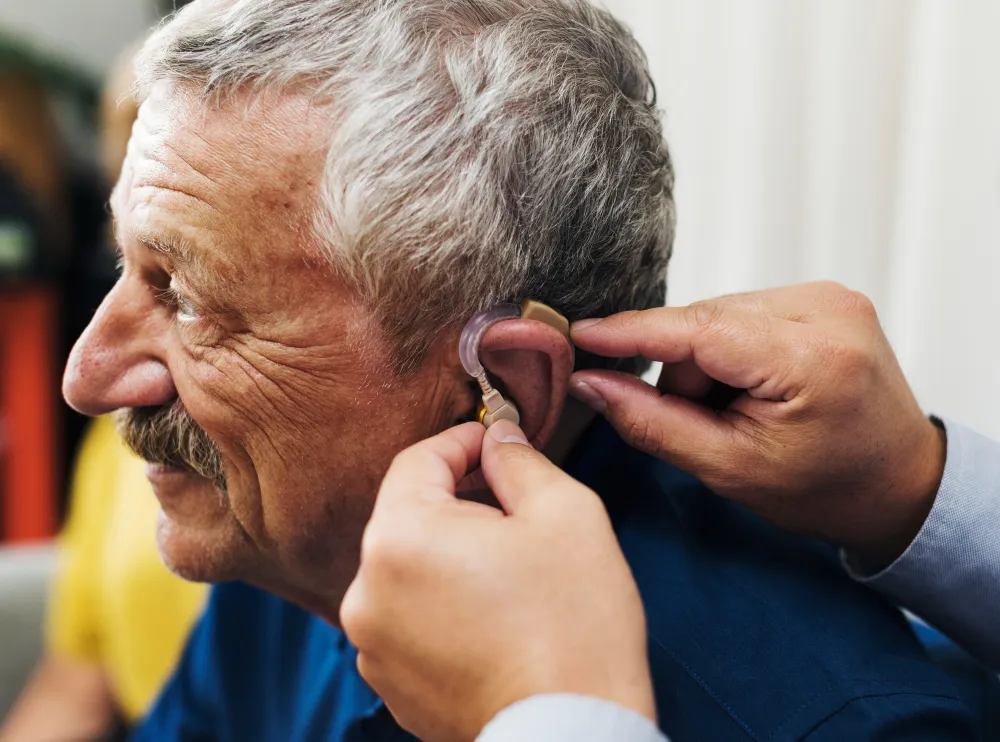 Elderly man wearing a hearing aid being adjusted by someone using their hands.
